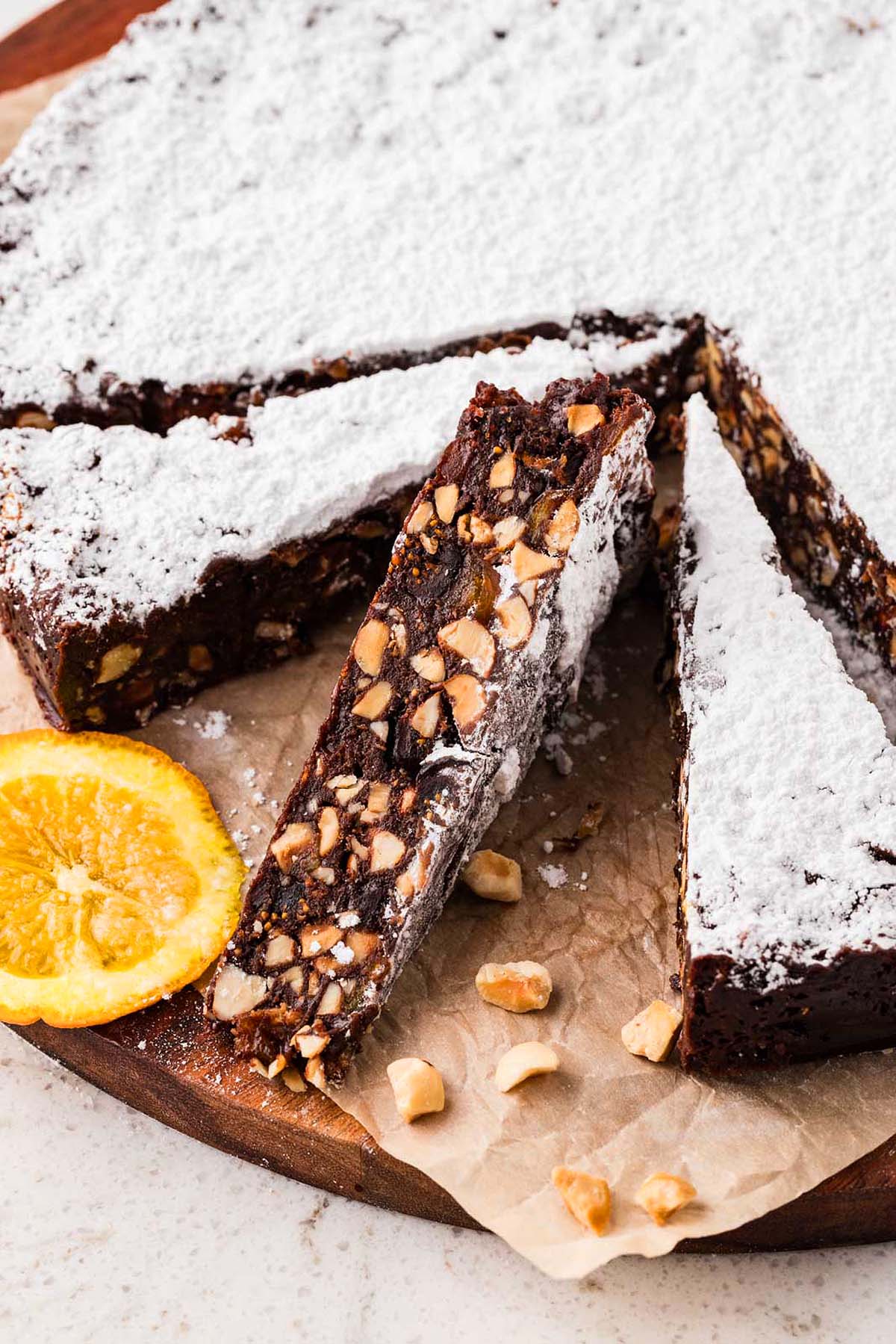Chocolate nut and fruit cake slices with powdered sugar on top, garnished with orange slices and chopped nuts.