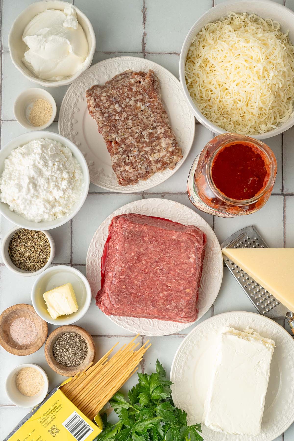 Million dollar spaghetti casserole ingredients: ground meat, cheeses, marinara, spices, butter, spaghetti, and fresh parsley on a counter.