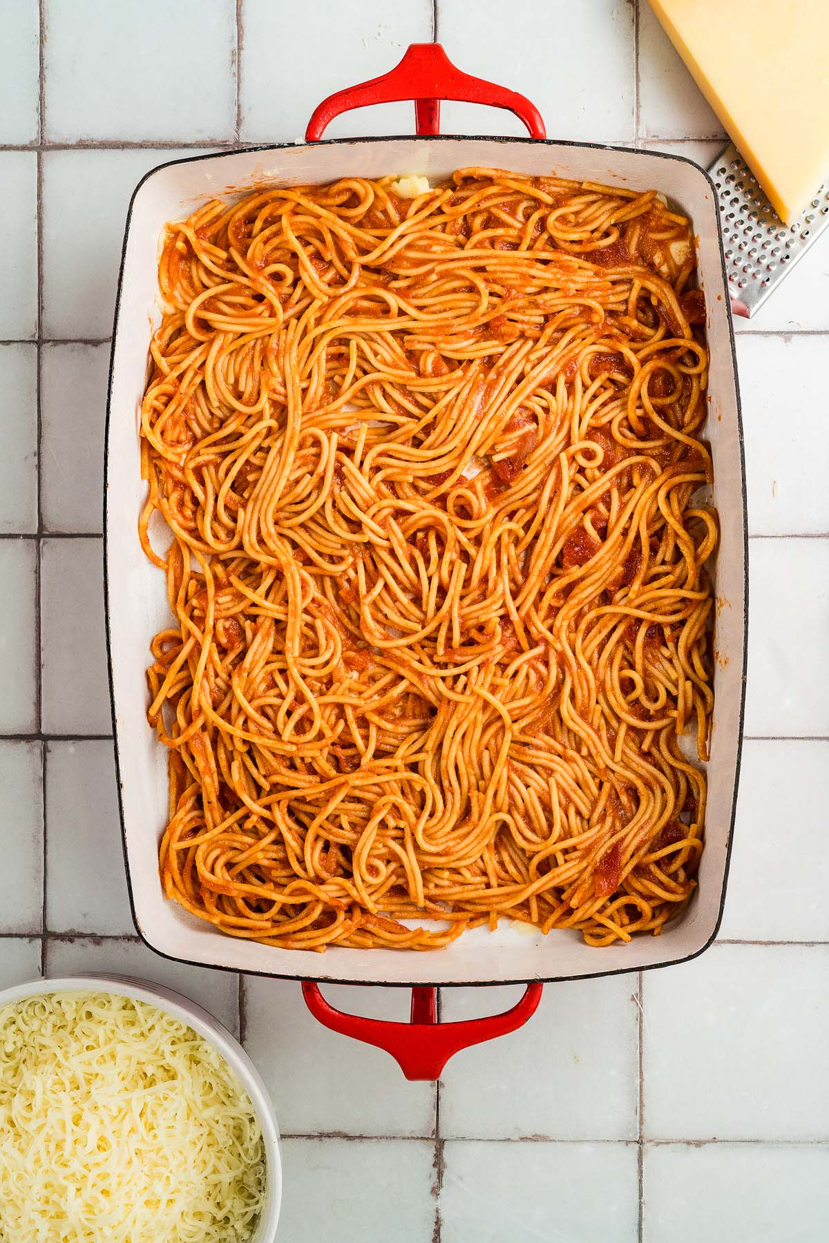 Baked spaghetti with tomato sauce in a red dish, next to shredded cheese and a block of cheese on a tiled surface.