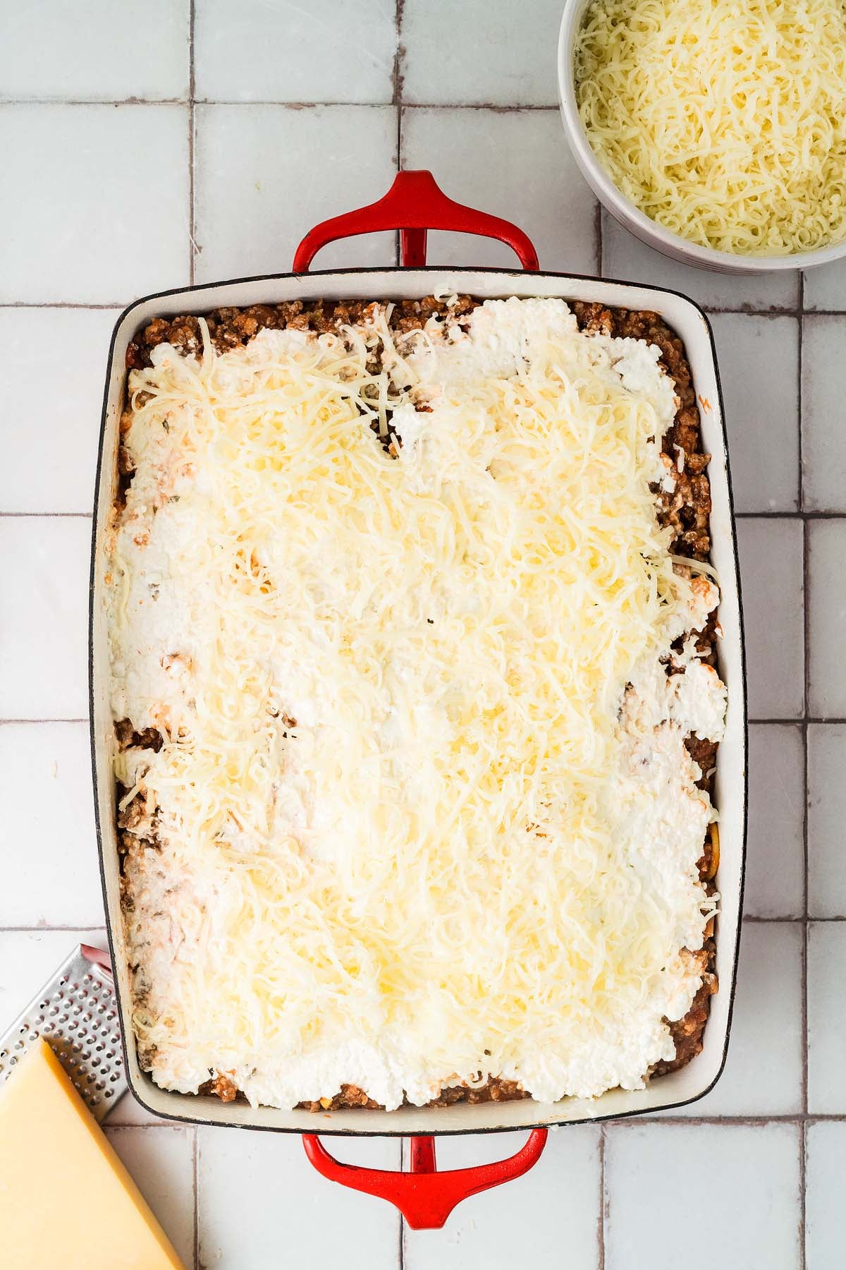 Rectangular baking dish with pasta, cheese on top, and a bowl of shredded cheese nearby.