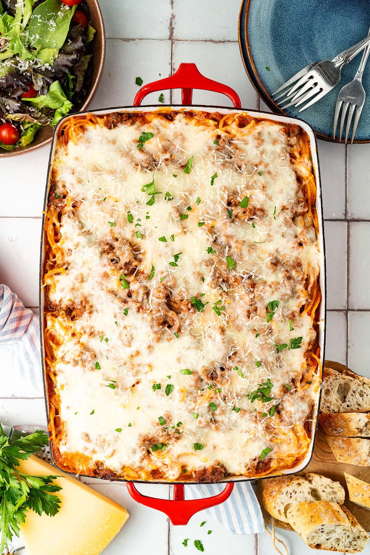 Baked spaghetti casserole with melted cheese, parsley on top, surrounded by salad, bread, and plates.