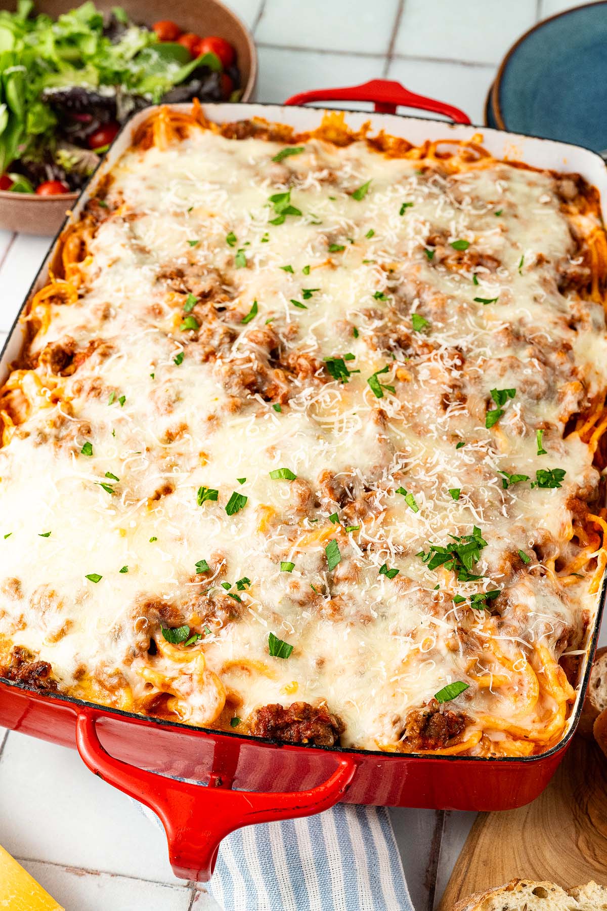 Baked spaghetti casserole topped with melted cheese and herbs in a red dish, with salad on the side.
