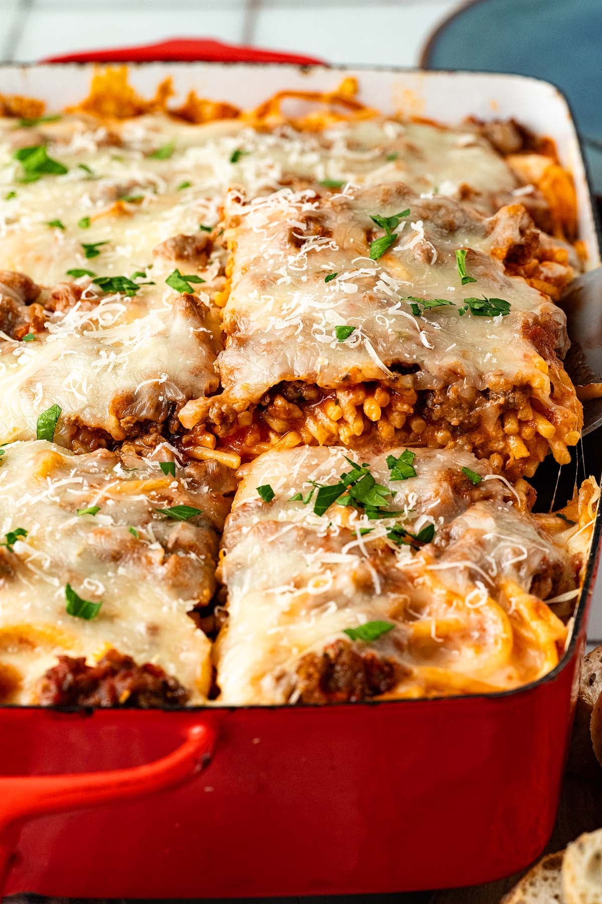 Baked casserole with melted cheese, ground meat, and pasta, topped with chopped parsley in a red dish.