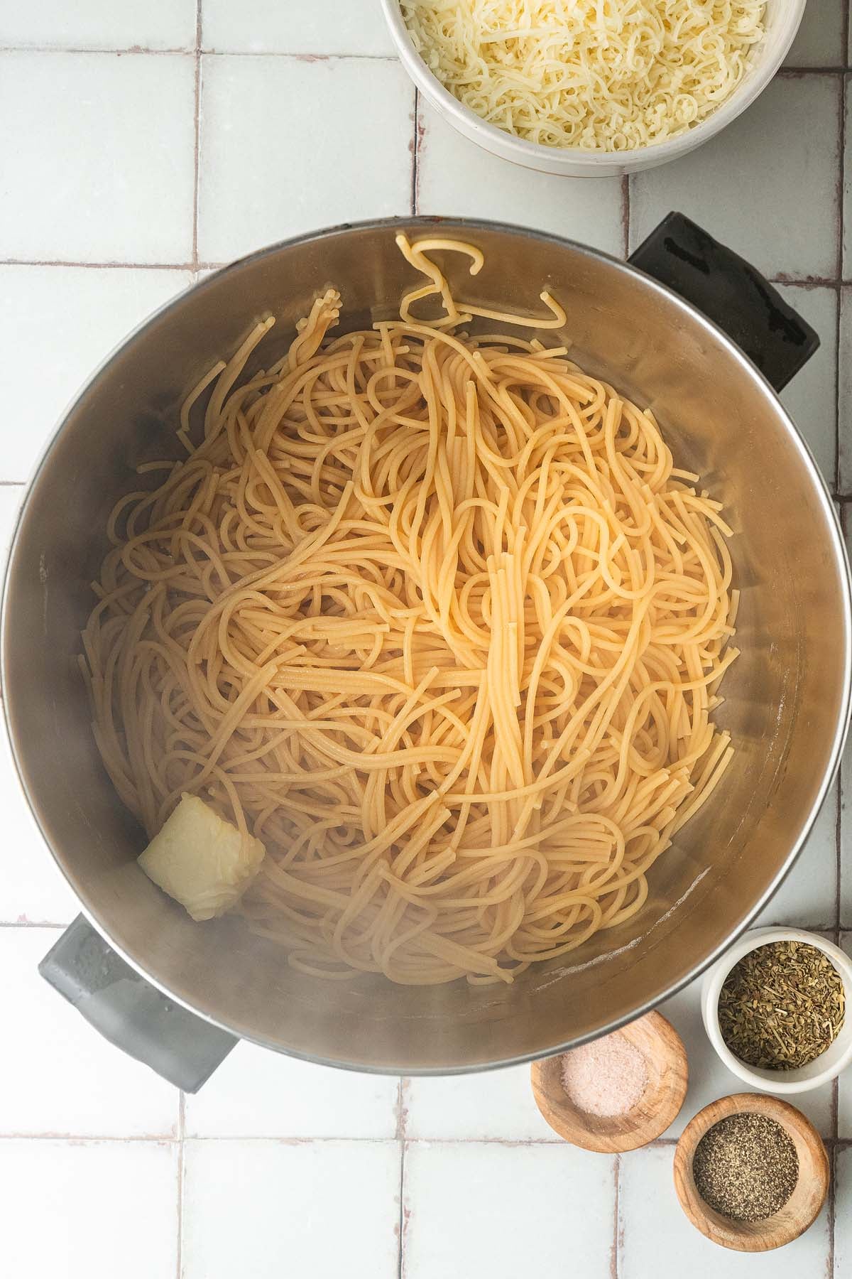 Cooked spaghetti in a pot with a pat of butter, surrounded by bowls of cheese, pepper, salt, and herbs.