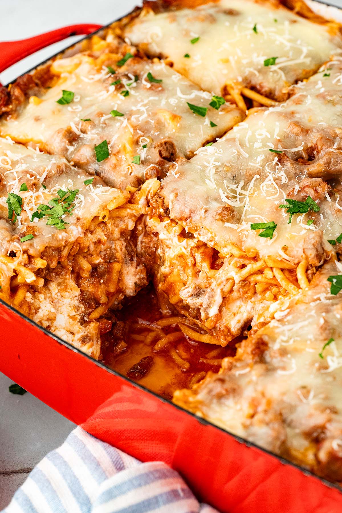 Baked spaghetti casserole with melted cheese, sliced in a red baking dish, garnished with chopped parsley.