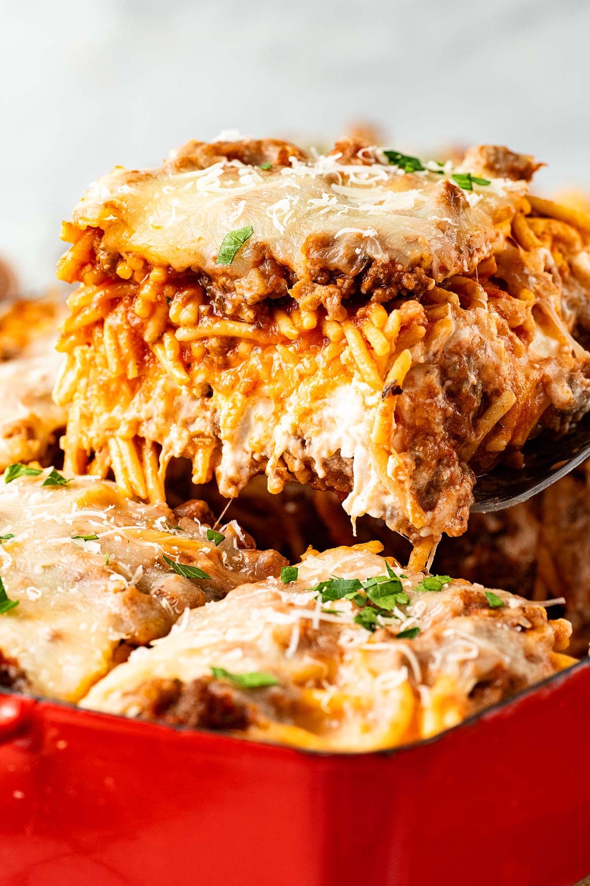 A cheesy baked spaghetti casserole is being served from a red dish, topped with parsley and grated cheese.