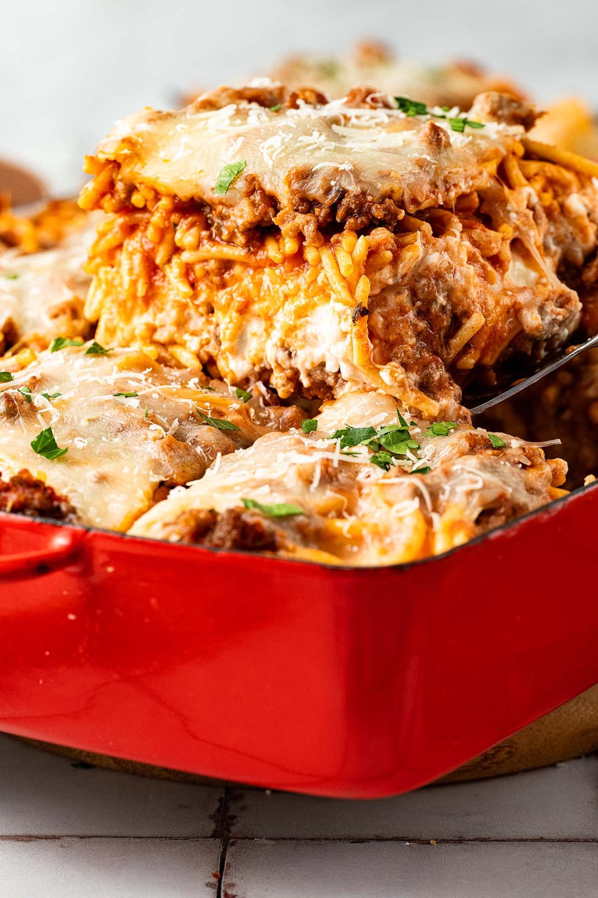 A close-up of cheesy million dollar spaghetti being served from a red baking dish, topped with herbs and grated cheese.