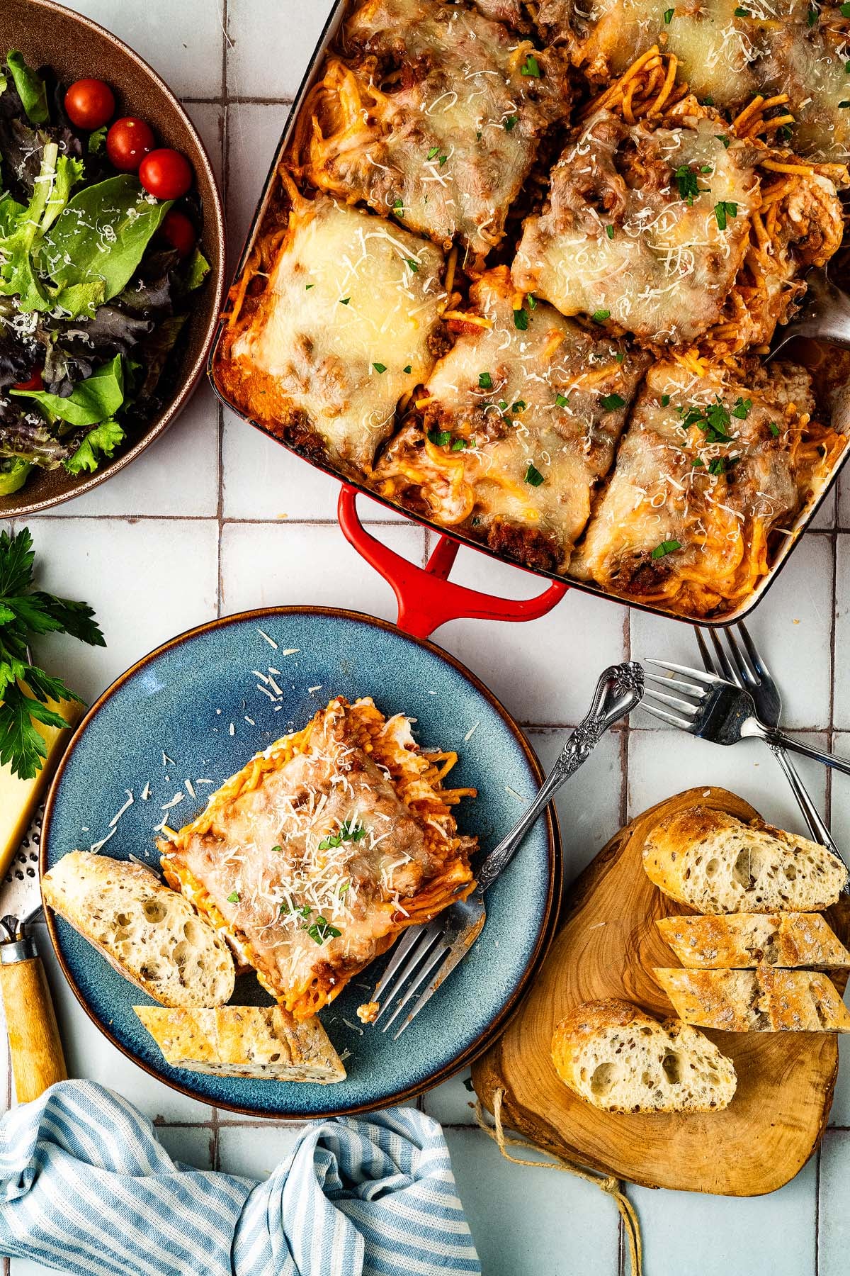 Baked spaghetti casserole served with salad and bread on a table, with a portion plated and garnished with cheese.