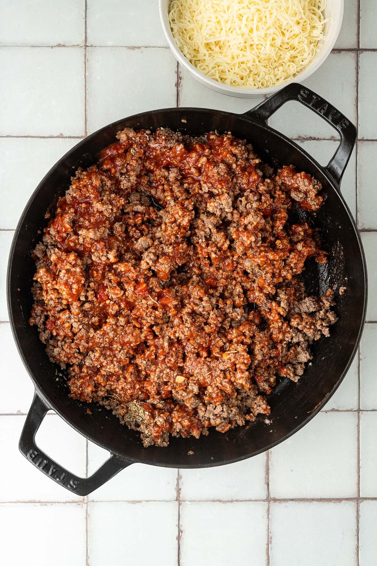 Cooked ground beef with tomato sauce in a black skillet, with a bowl of shredded cheese nearby.