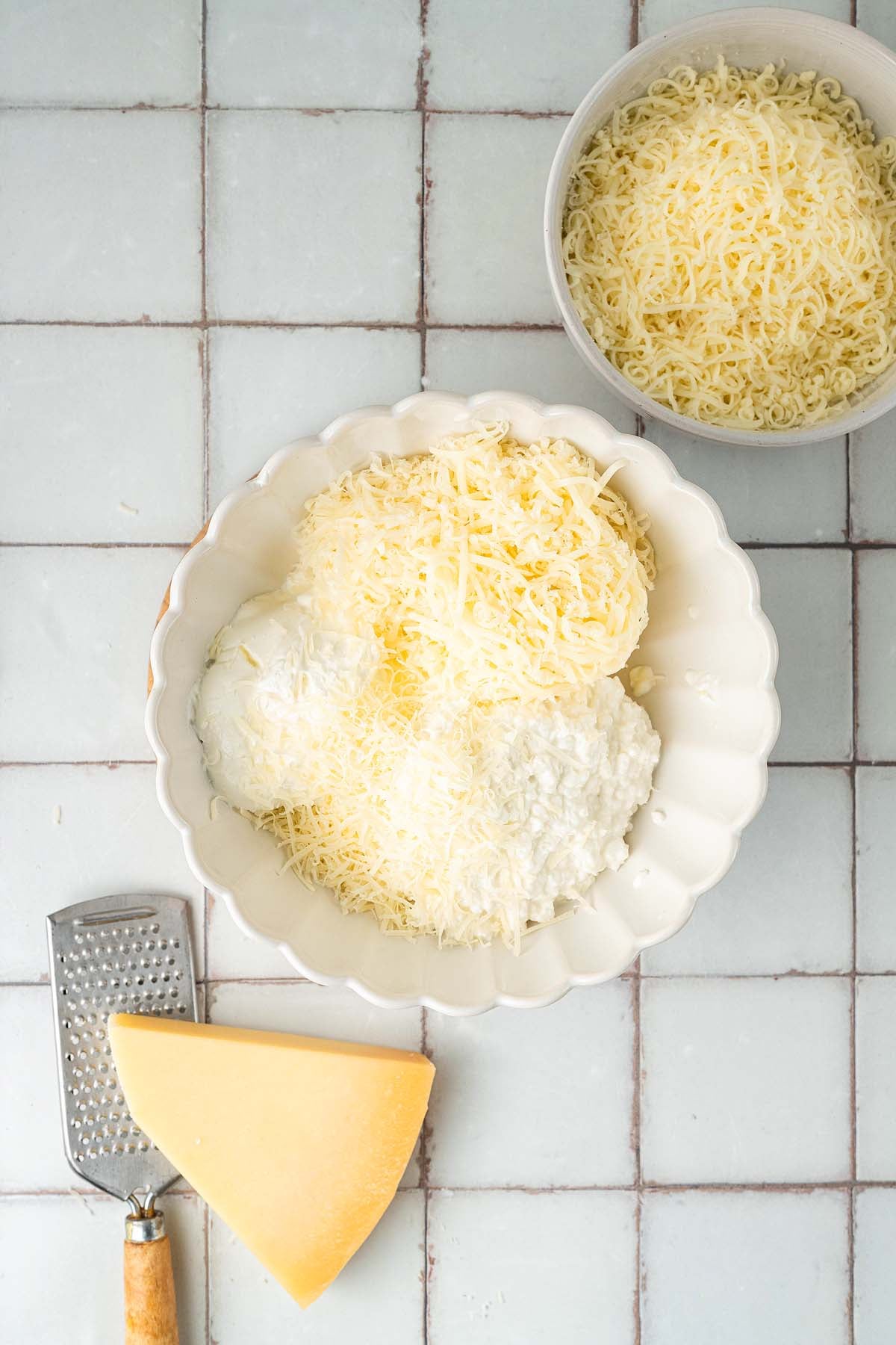 Two bowls of shredded cheese, one with cottage cheese, next to a cheese wedge and grater on a tiled surface.