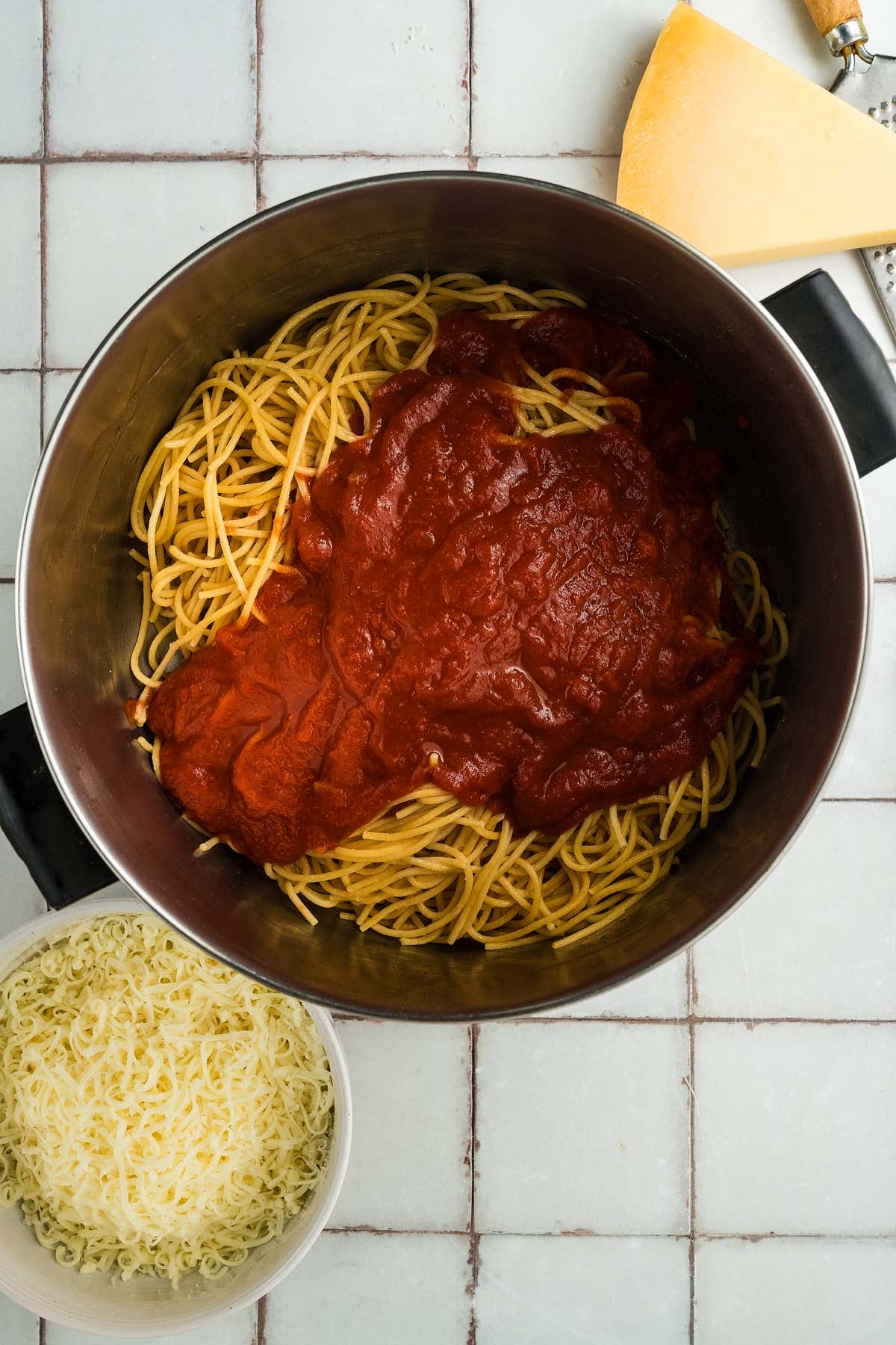 Cooked spaghetti in a pot topped with tomato sauce, next to shredded cheese and a wedge of cheese.