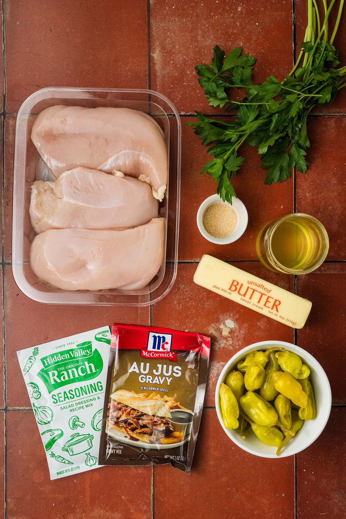 Raw chicken breasts, parsley, butter, ranch seasoning, au jus, pepperoncini, garlic powder, and broth on tile.