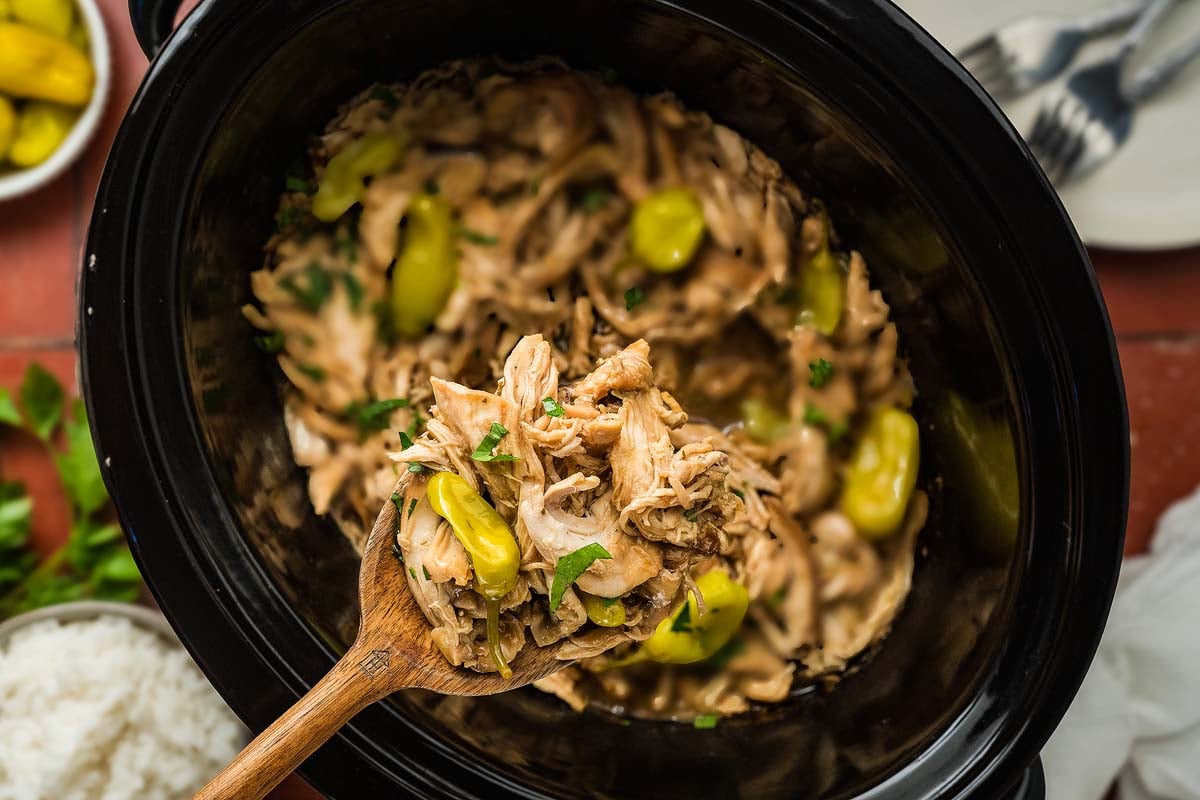 Shredded chicken and peppers in a slow cooker, served with a wooden spoon, garnished with fresh herbs.