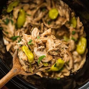 Shredded chicken with peppers and herbs in a slow cooker, served with a wooden spoon.