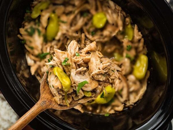 Shredded chicken with peppers and herbs in a slow cooker, served with a wooden spoon.
