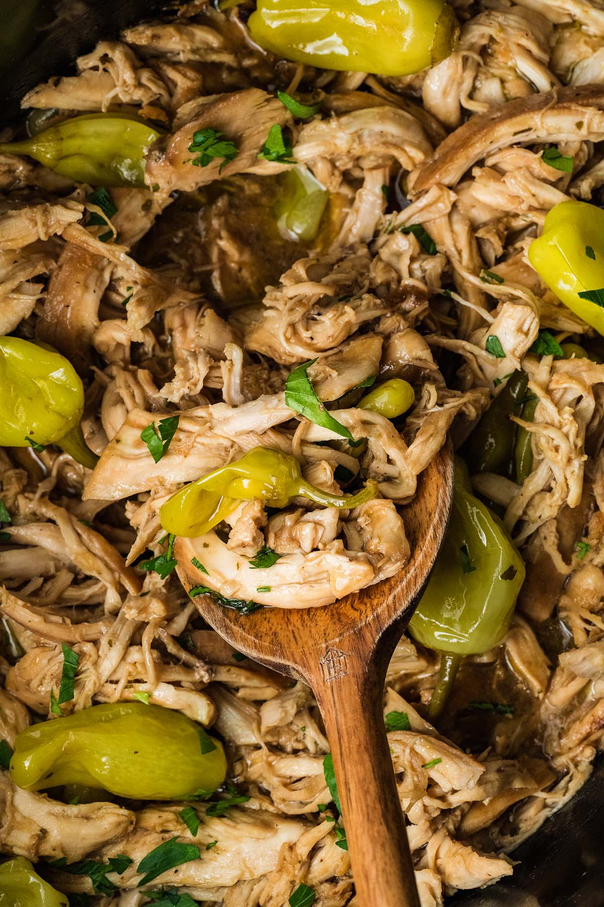 Shredded chicken with pepperoncini and herbs in a pot, stirred with a wooden spoon.