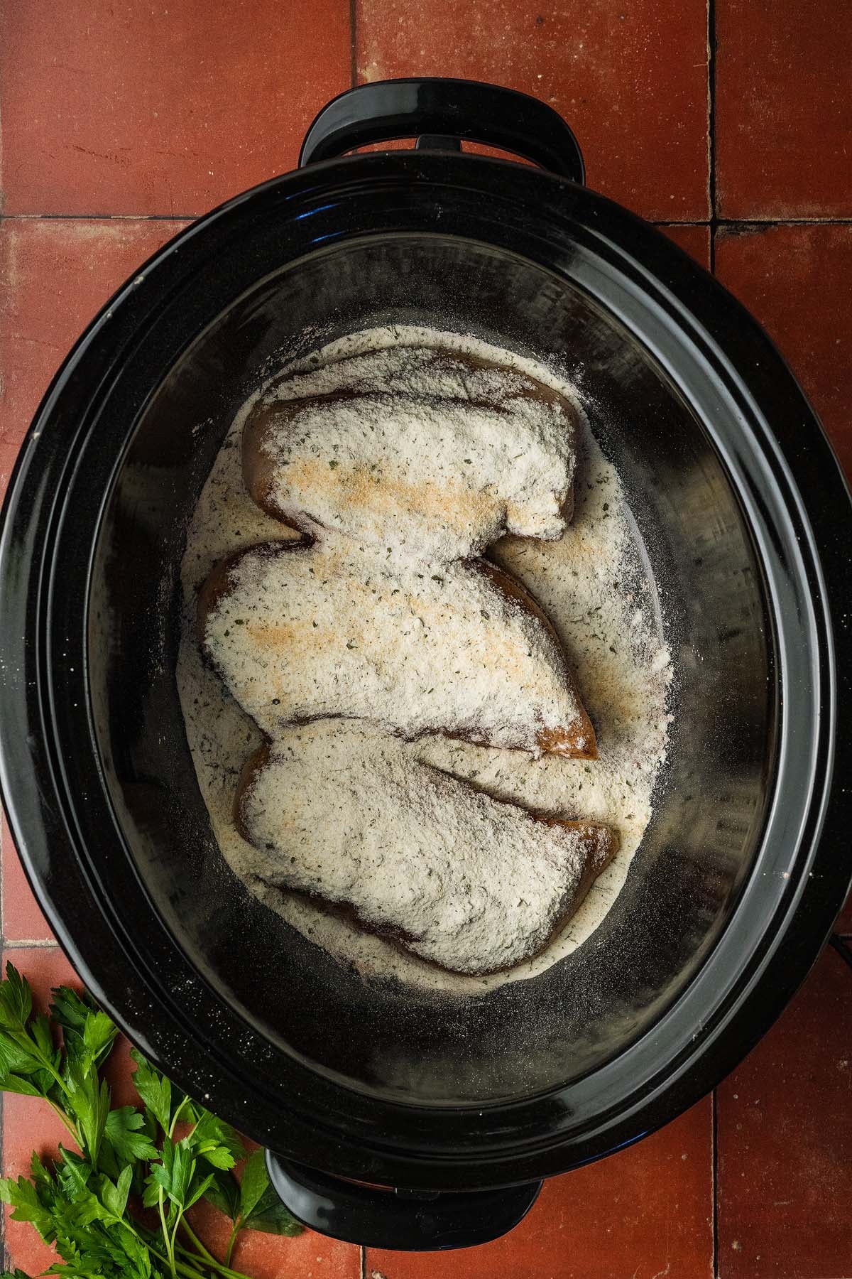 Three chicken breasts in a slow cooker, on a red tiled counter with parsley nearby.