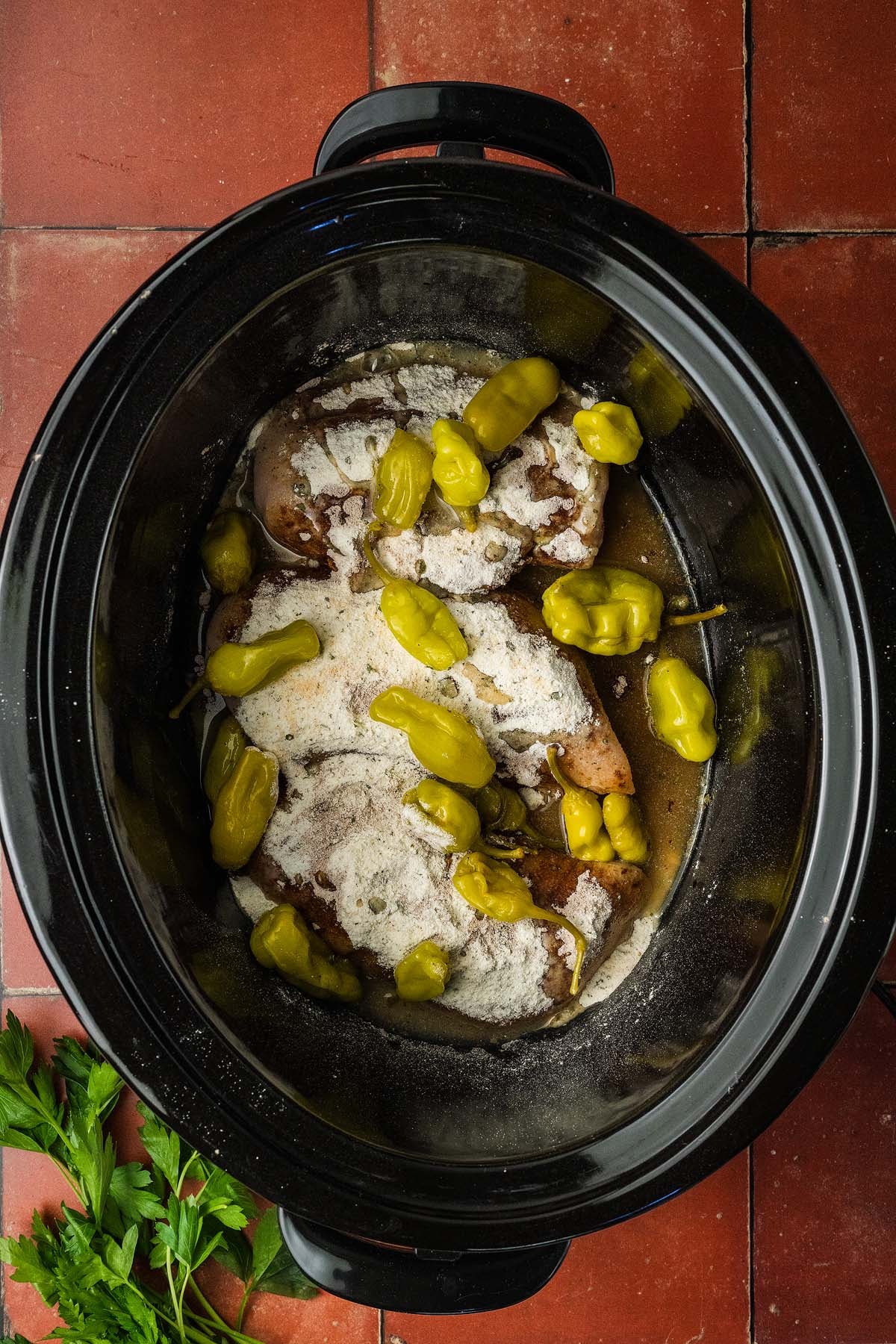 Raw chicken, seasoning, and pepperoncini peppers in a slow cooker on a red tiled surface, parsley nearby.