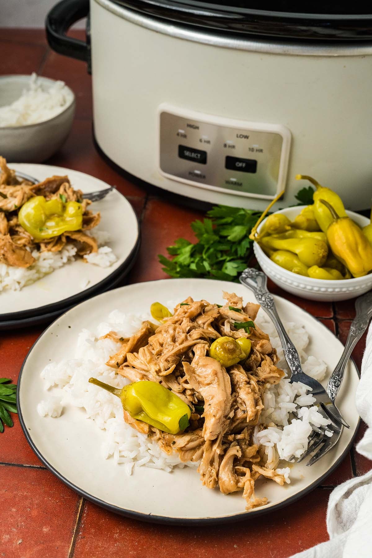 Shredded chicken with peppers served over rice on plates, next to a slow cooker and a bowl of peppers.