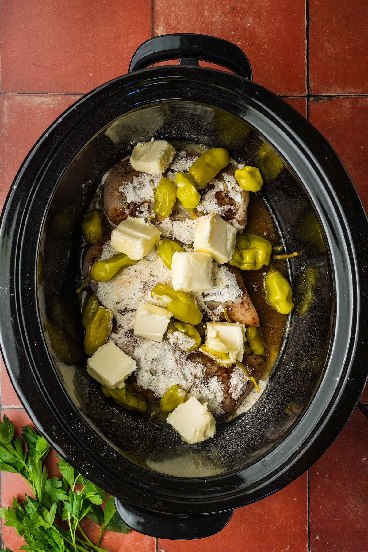 Raw chicken, pepperoncini, butter, and seasoning in a slow cooker, on a red tiled countertop with parsley.