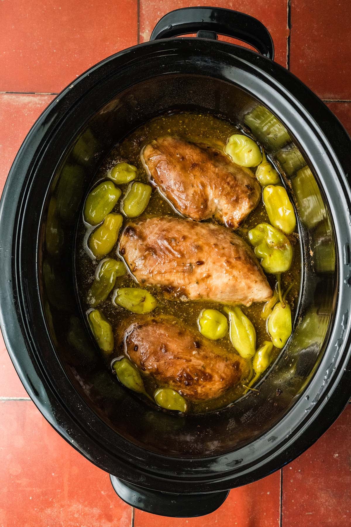 Three cooked chicken breasts and peppers in sauce inside a black slow cooker on a red tiled surface.