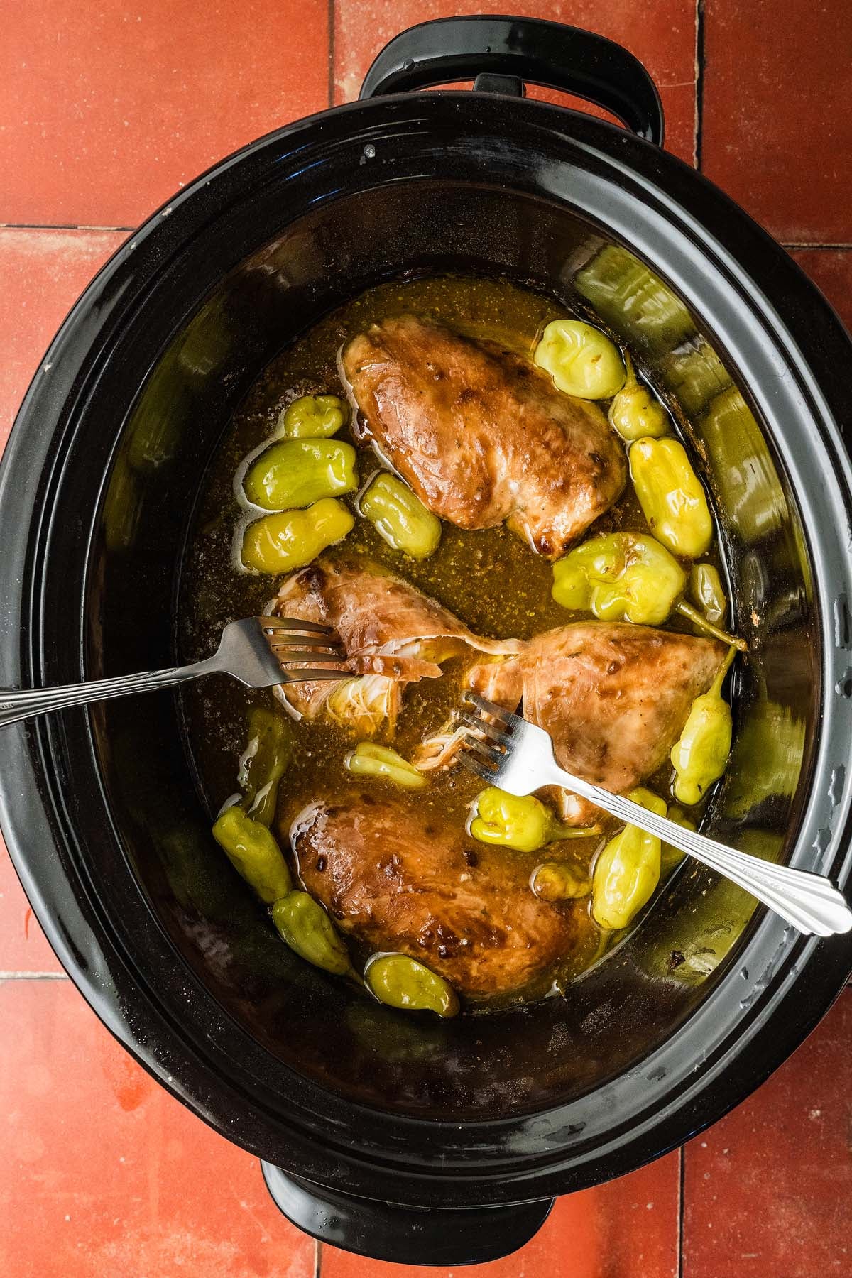 Chicken breasts and peppers in a slow cooker with two forks, ready to shred, on a red tiled surface.