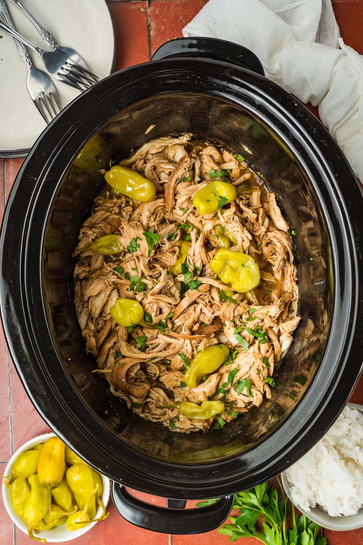Shredded chicken with pepperoncini peppers in a slow cooker, garnished with herbs, on a red tiled surface.