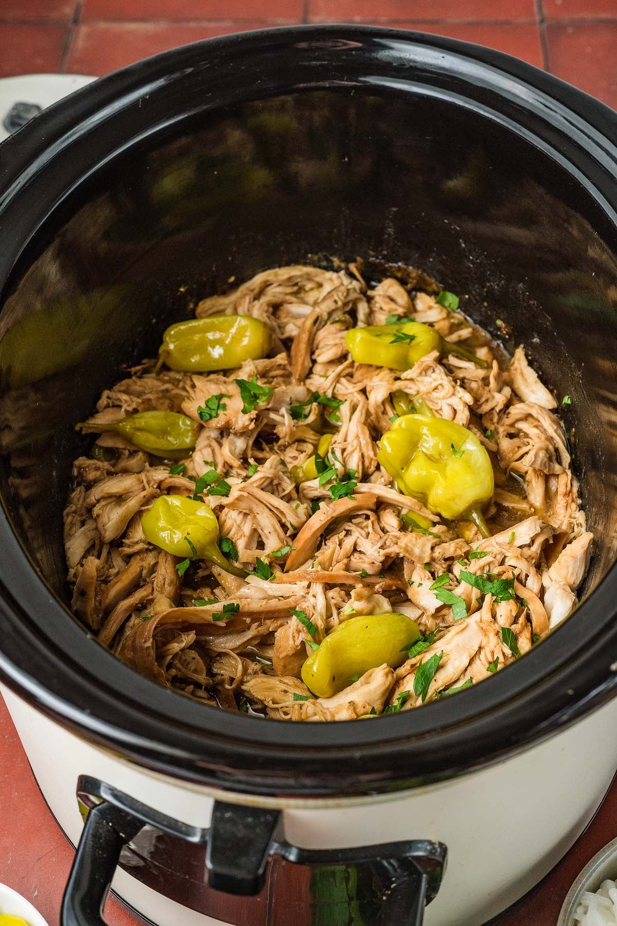 Shredded chicken with pepperoncini peppers and herbs in a black slow cooker on a red tiled countertop.