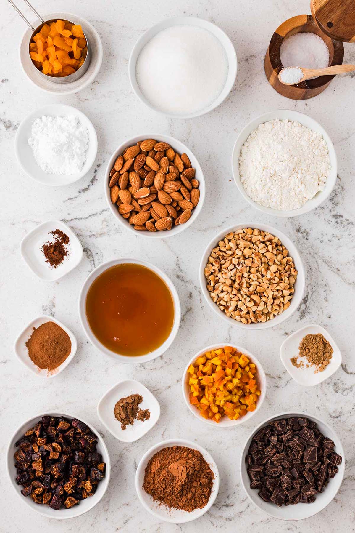 Bowls of dried fruit, nuts, chocolate, honey, spices, flour, and sugar arranged on a marble countertop.