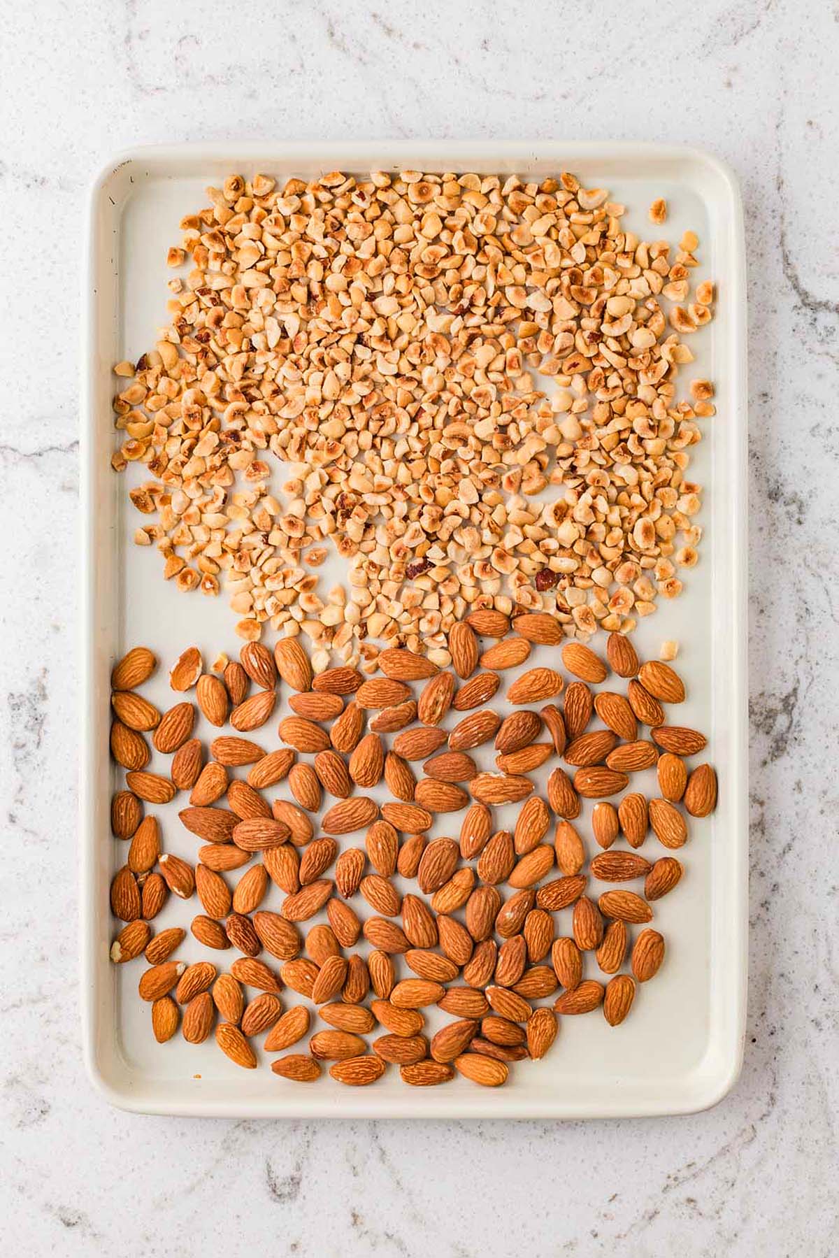 A tray with chopped nuts on top and whole almonds on the bottom, on a marble surface.