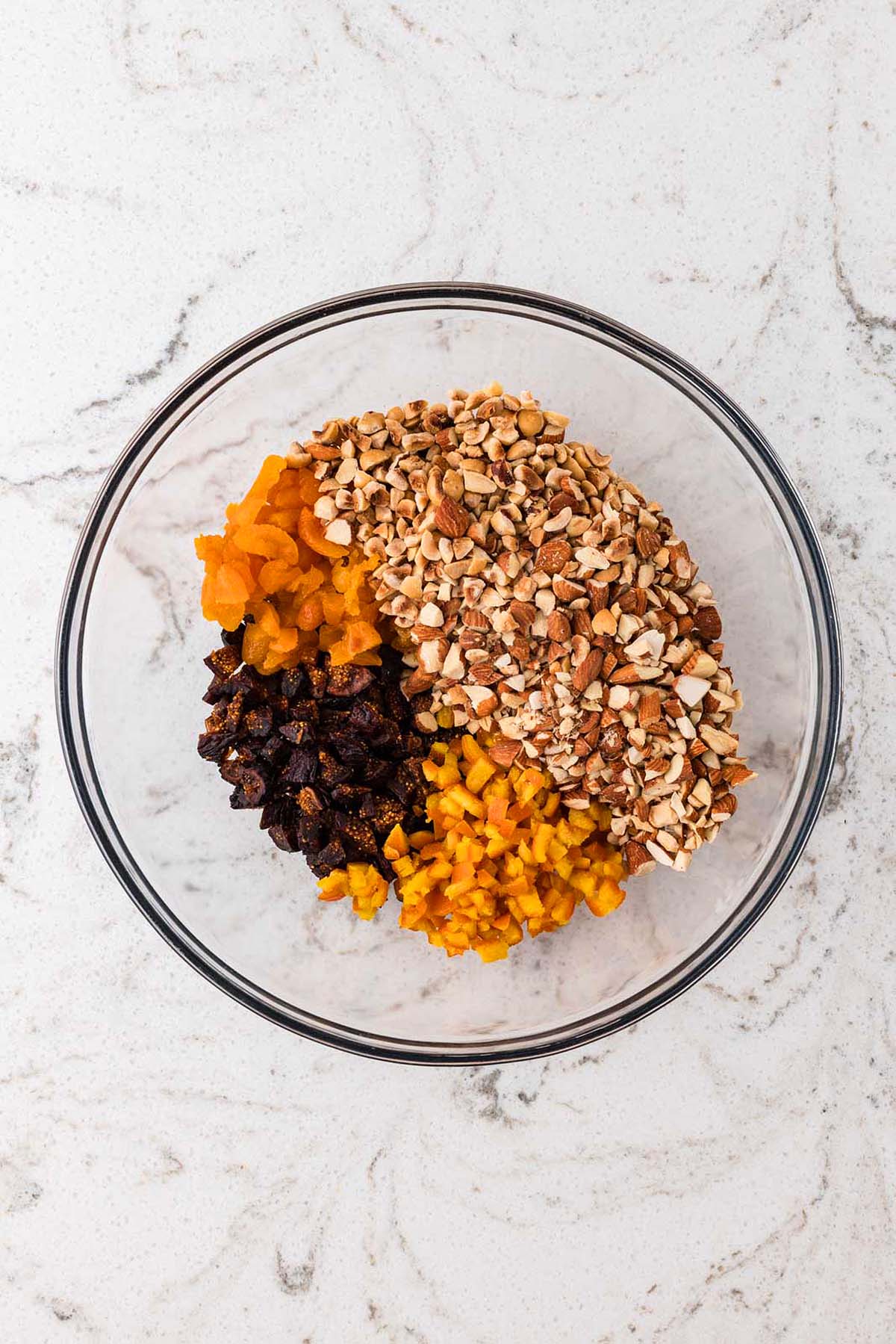 Glass bowl with chopped dried fruit and mixed nuts on a white marble surface.