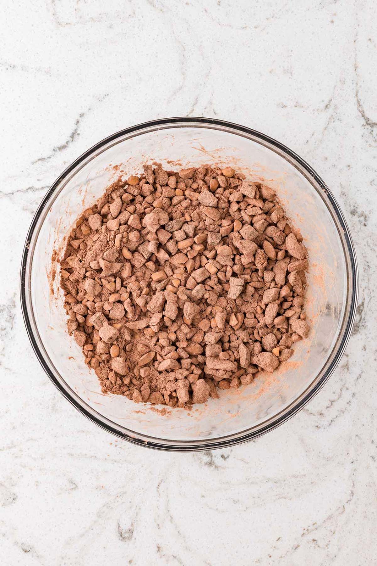 A glass bowl filled with a chunky cocoa powder mixture sits on a white marble countertop.
