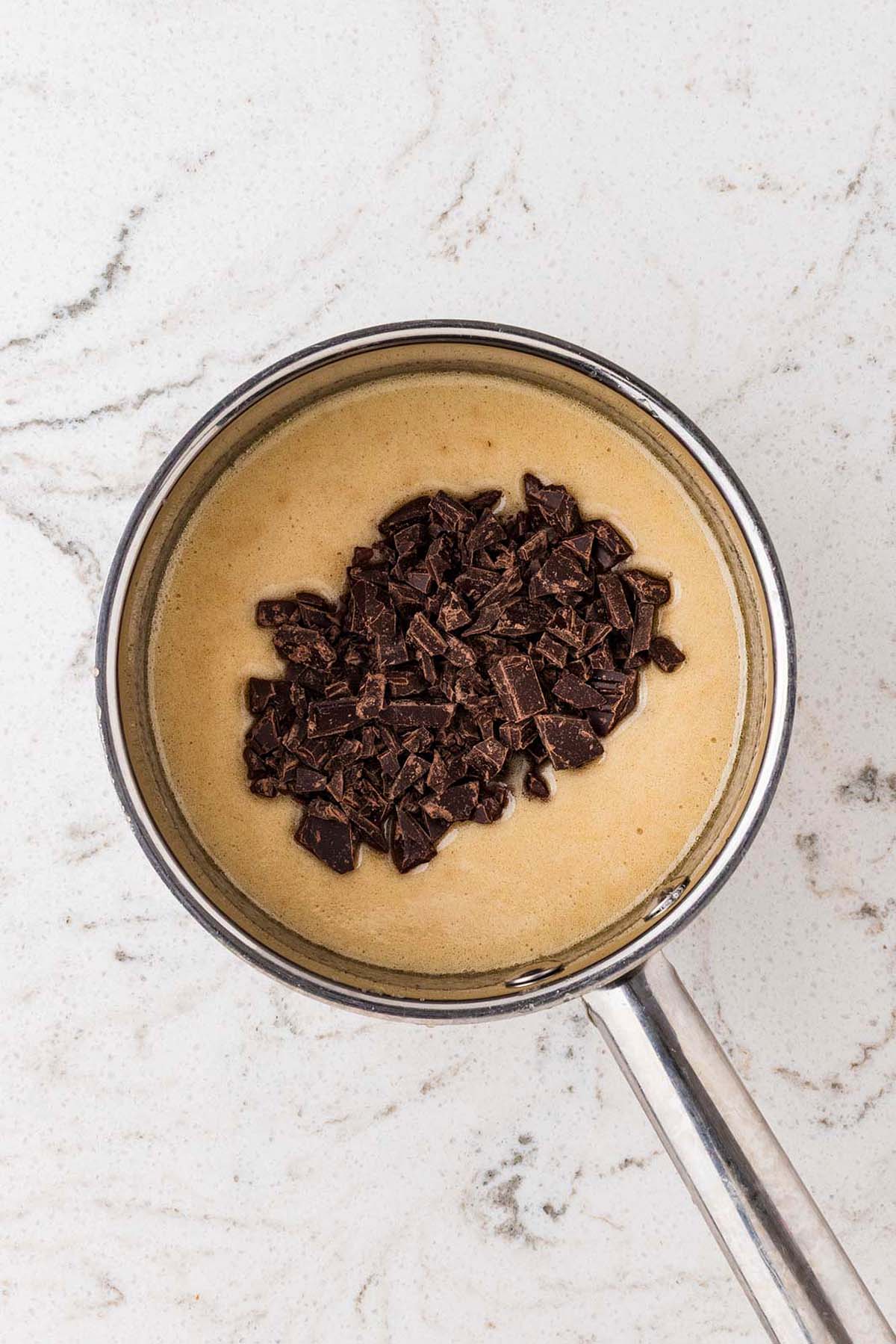 Chopped chocolate sits atop creamy mixture in a saucepan on a white marble countertop.