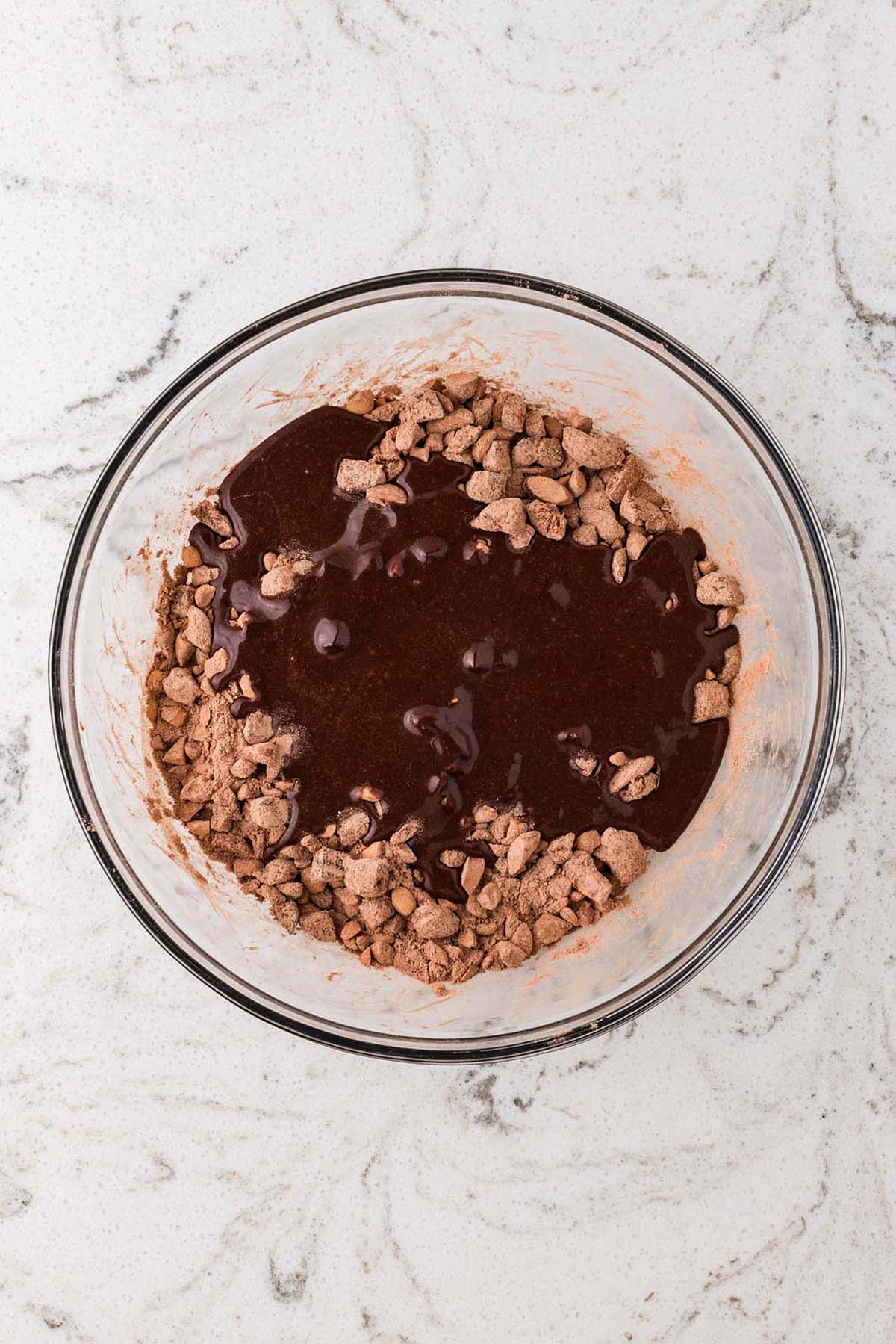 A glass bowl with cocoa powder and melted chocolate mixing on a white marble countertop.