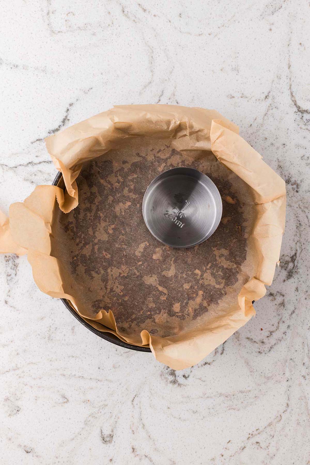 A round baking pan lined with parchment paper, with a metal measuring cup inside, on a marble surface.