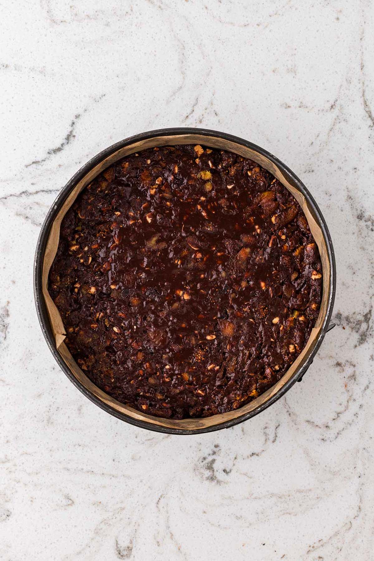 Round baking pan with chocolate and a fruit and nut mixture on white marbled surface, top view.