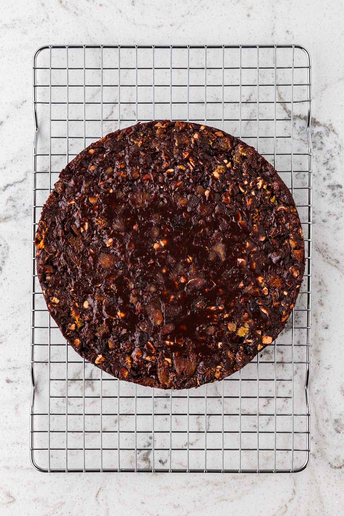 A round chocolate cake with fruit and nuts cooling on a wire rack over a marble surface.