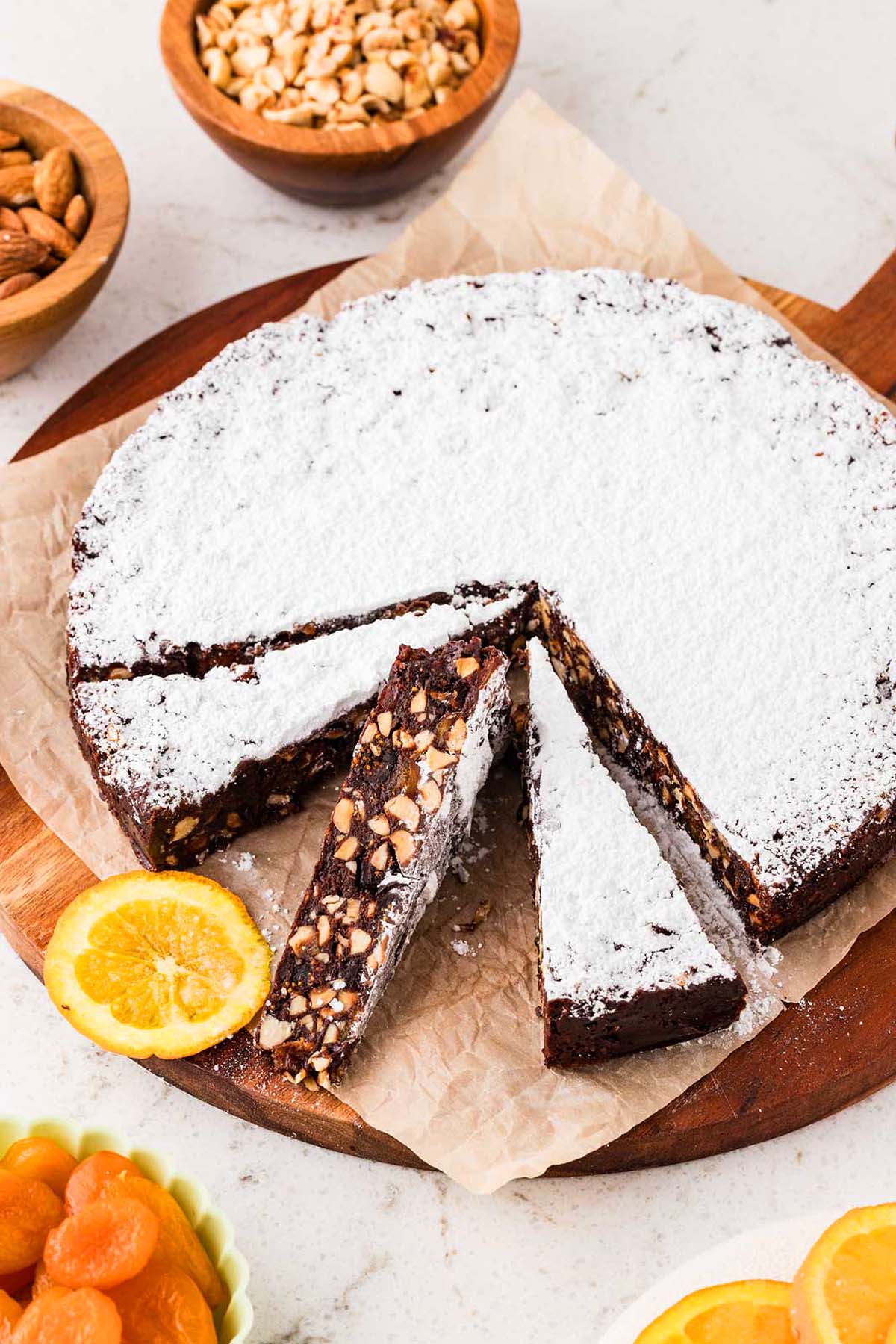 Round fruit and nut cake dusted with powdered sugar, with two slices cut, surrounded by nuts and dried fruit.