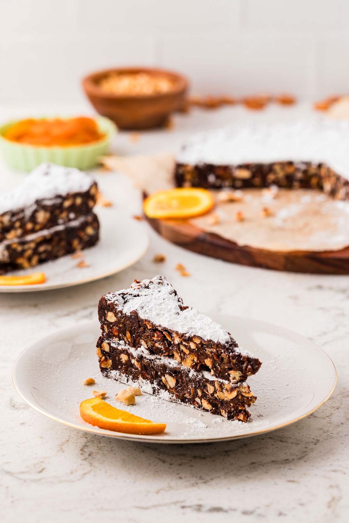 Two slices of chocolate nut cake with powdered sugar on a plate, garnished with an orange slice.