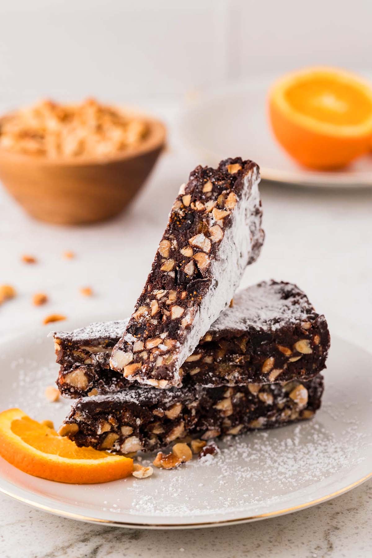 Three nut-studded chocolate panforte slices stacked on a plate, dusted with powdered sugar, next to an orange slice.