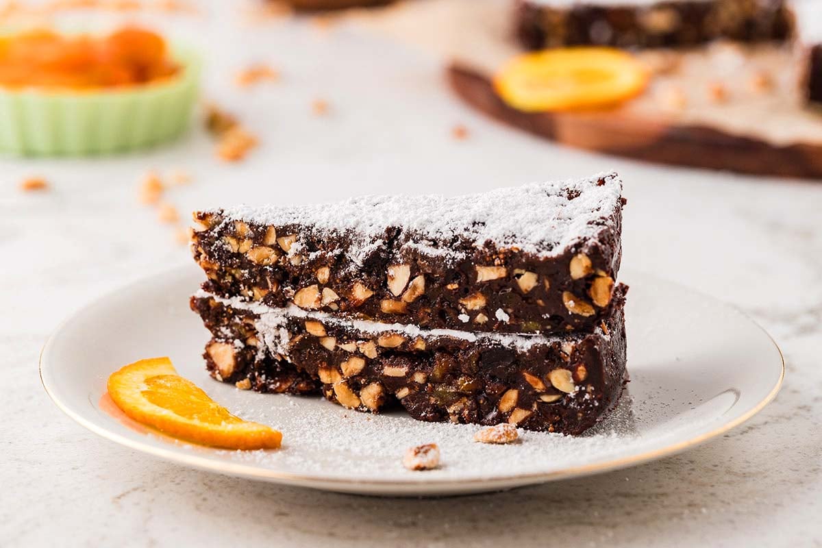 Two slices of nutty chocolate cake dusted with powdered sugar, served with an orange slice on a white plate.