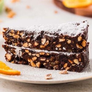 Two slices of chocolate panforte cake dusted with powdered sugar, served with an orange slice on a white plate.