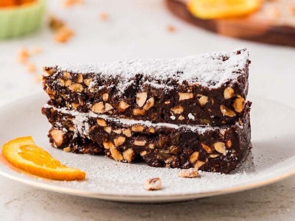 Two slices of chocolate panforte cake dusted with powdered sugar, served with an orange slice on a white plate.