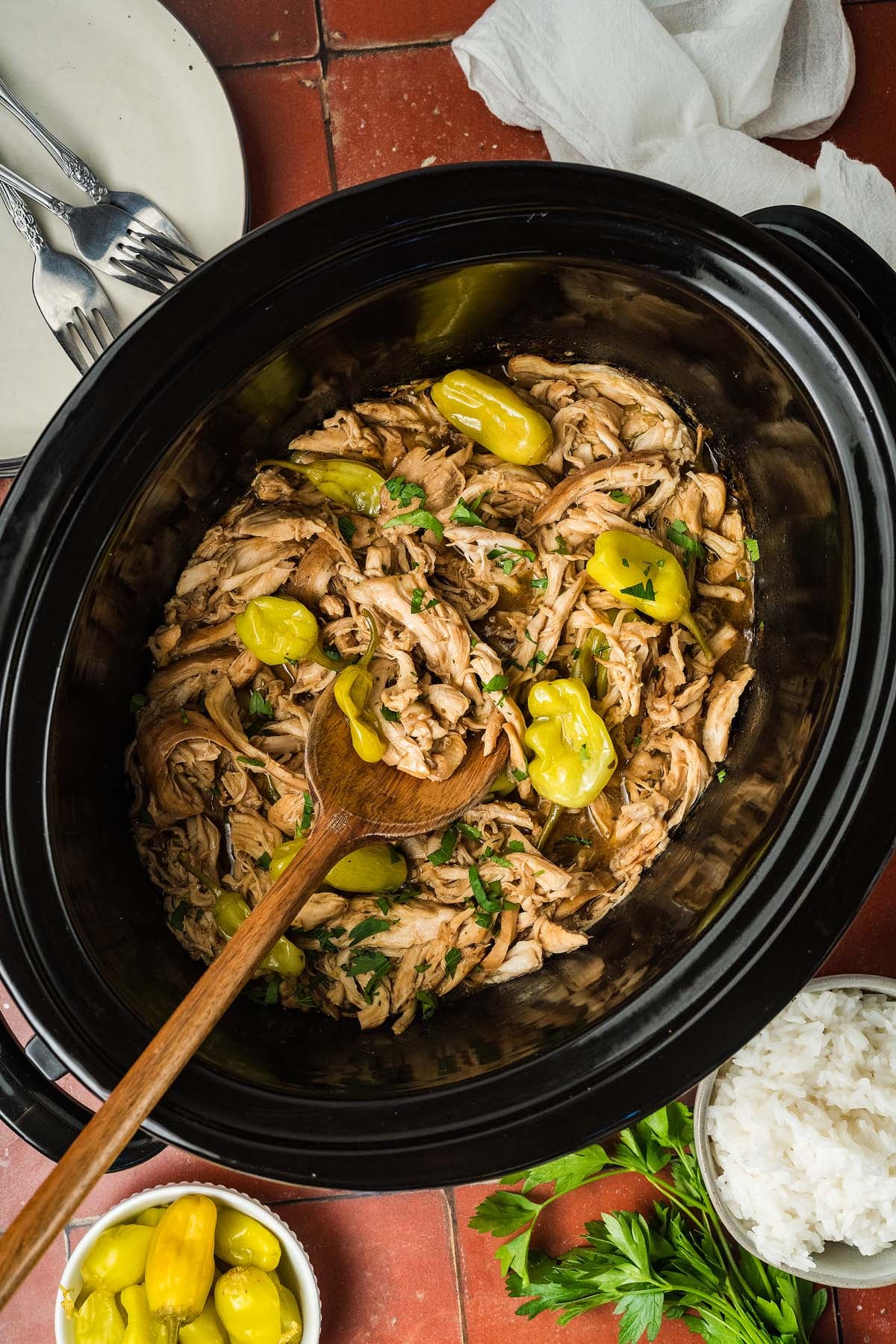 Shredded chicken with pepperoncini in a slow cooker, garnished with herbs, next to rice and parsley.