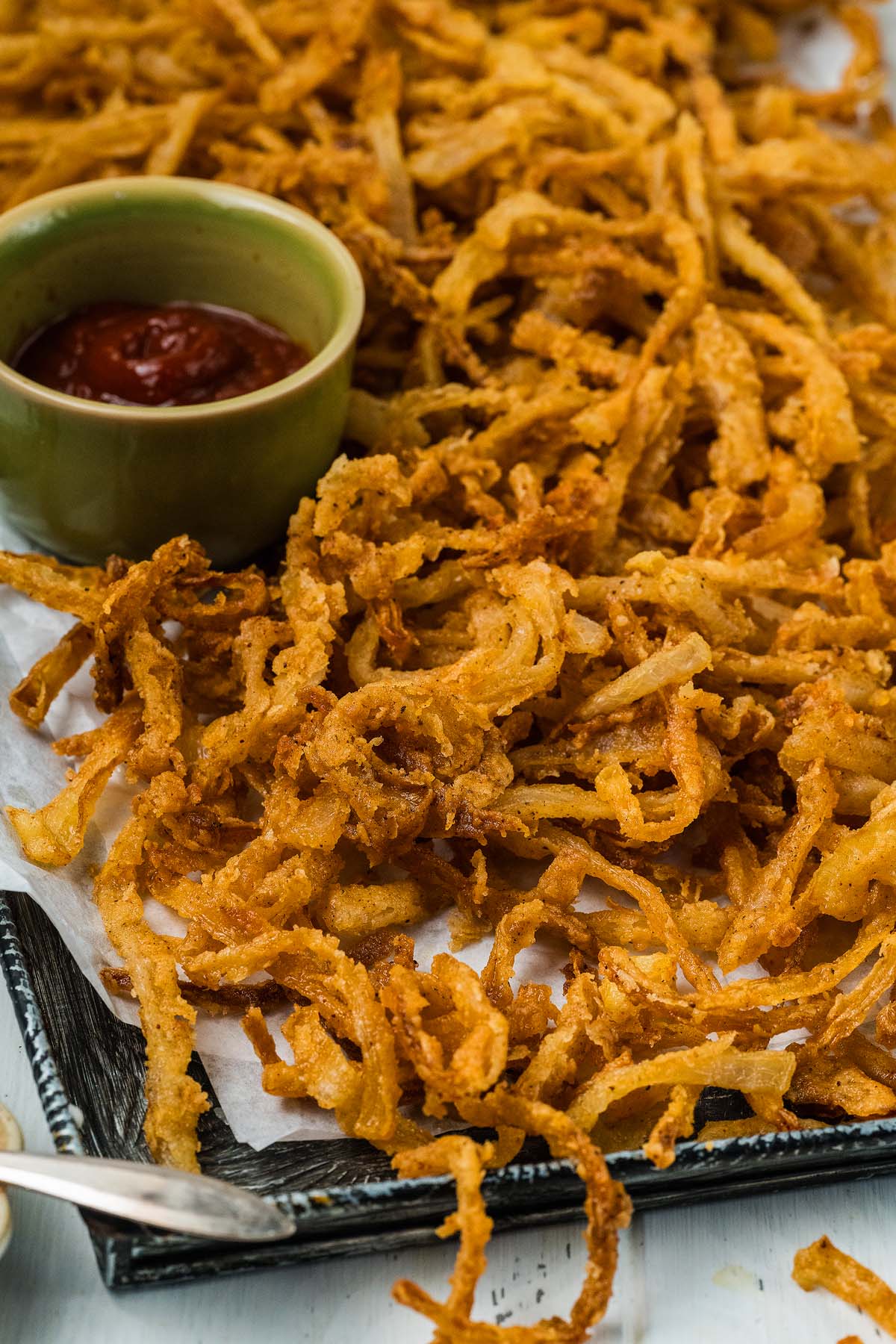 Crispy onion strings are piled high on a tray, served alongside a small green bowl filled with vibrant red dipping sauce.