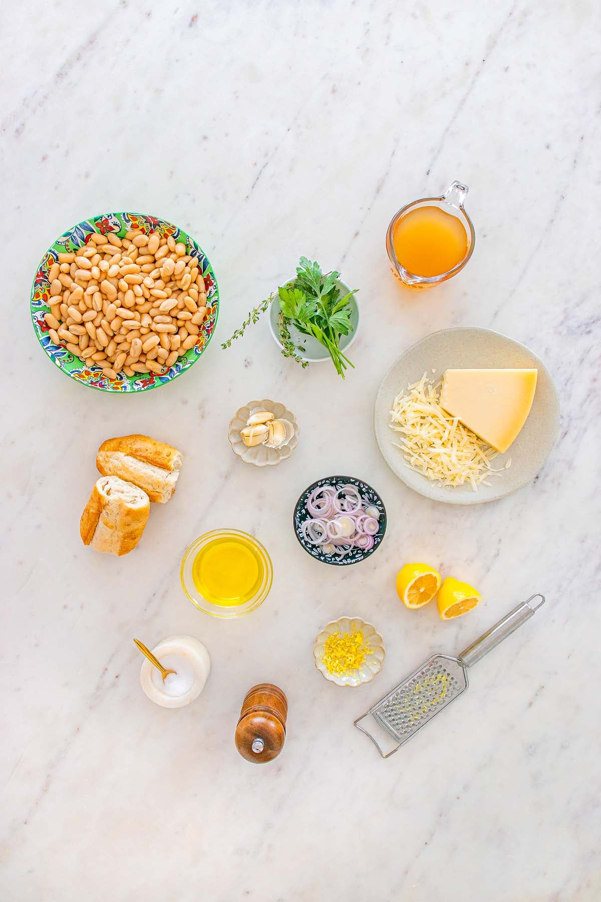 Ingredients on a marble surface: Tuscan white beans, cheese, bread, herbs, oil, broth, shallots, lemon, garlic, salt, pepper.