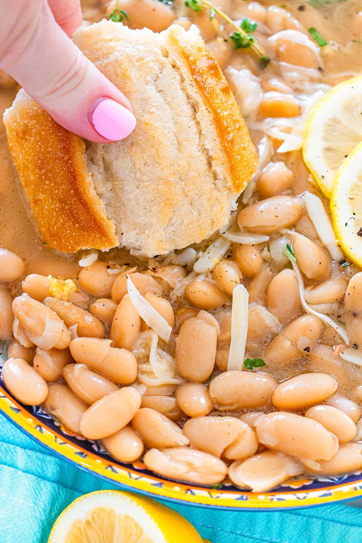 Hand dipping bread into a bowl of Tuscan white beans with onions, lemon slices, and herbs in broth.