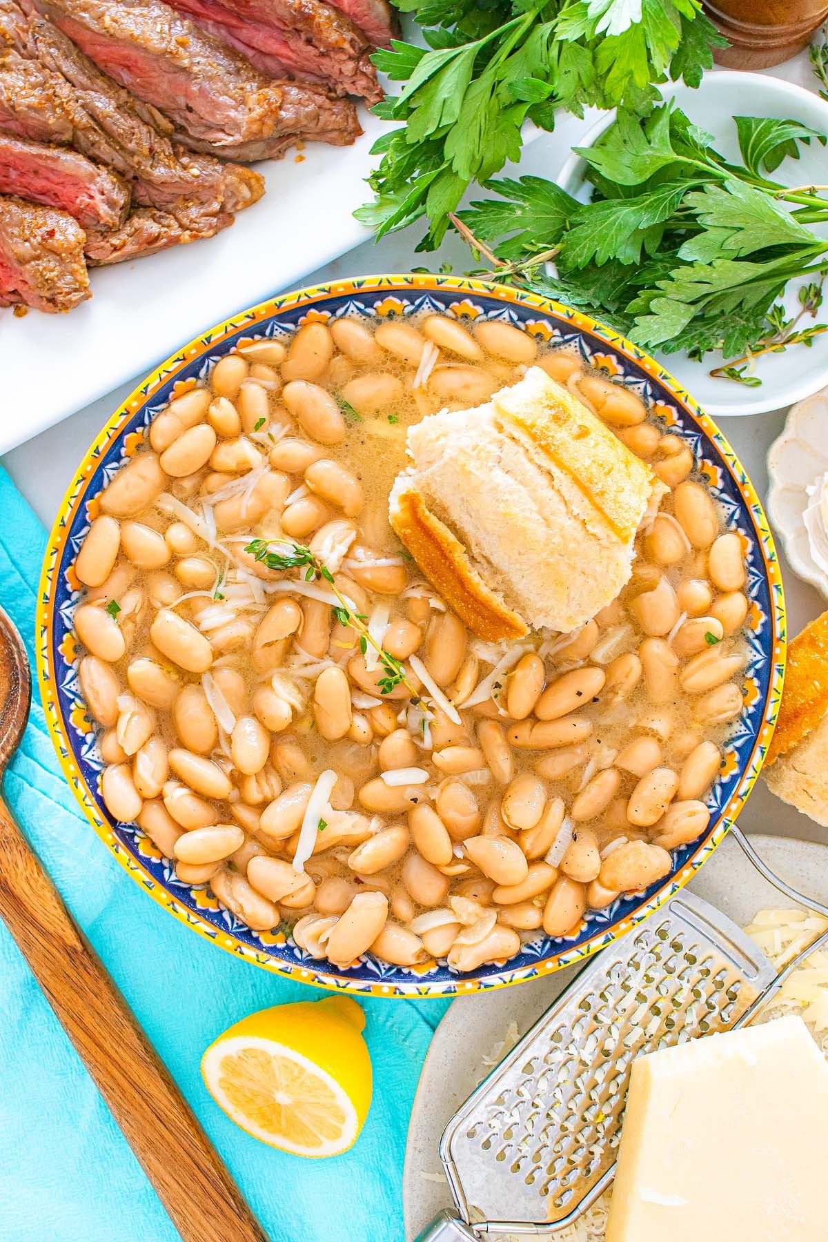 A bowl of hearty white bean soup made with Tuscan white beans and fresh herbs is served alongside crusty bread and surrounded by parsley, steak, cheese, and a bright lemon wedge.