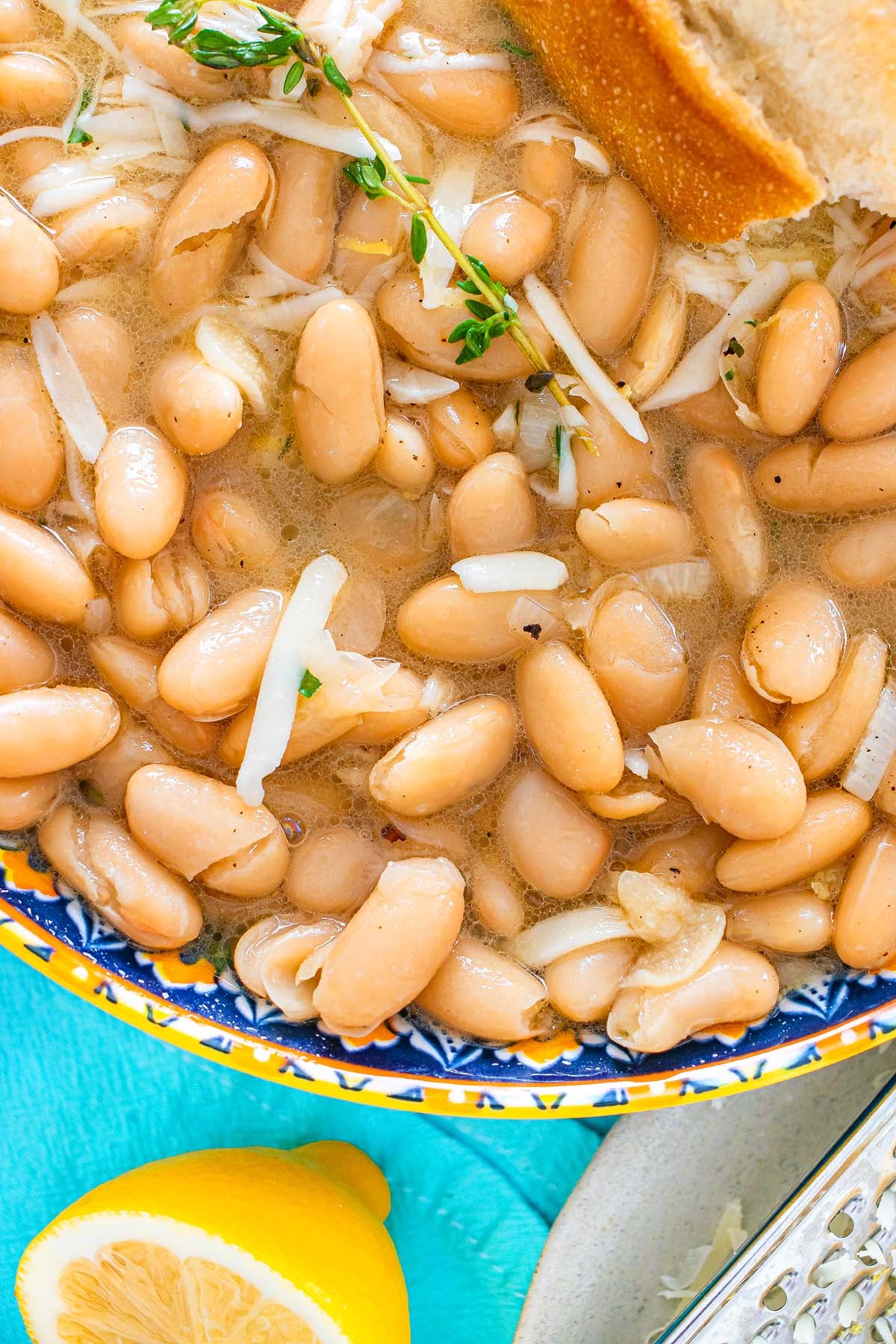 Tuscan white beans in broth with shredded cheese, fresh thyme, and bread, served with a lemon wedge on the side.