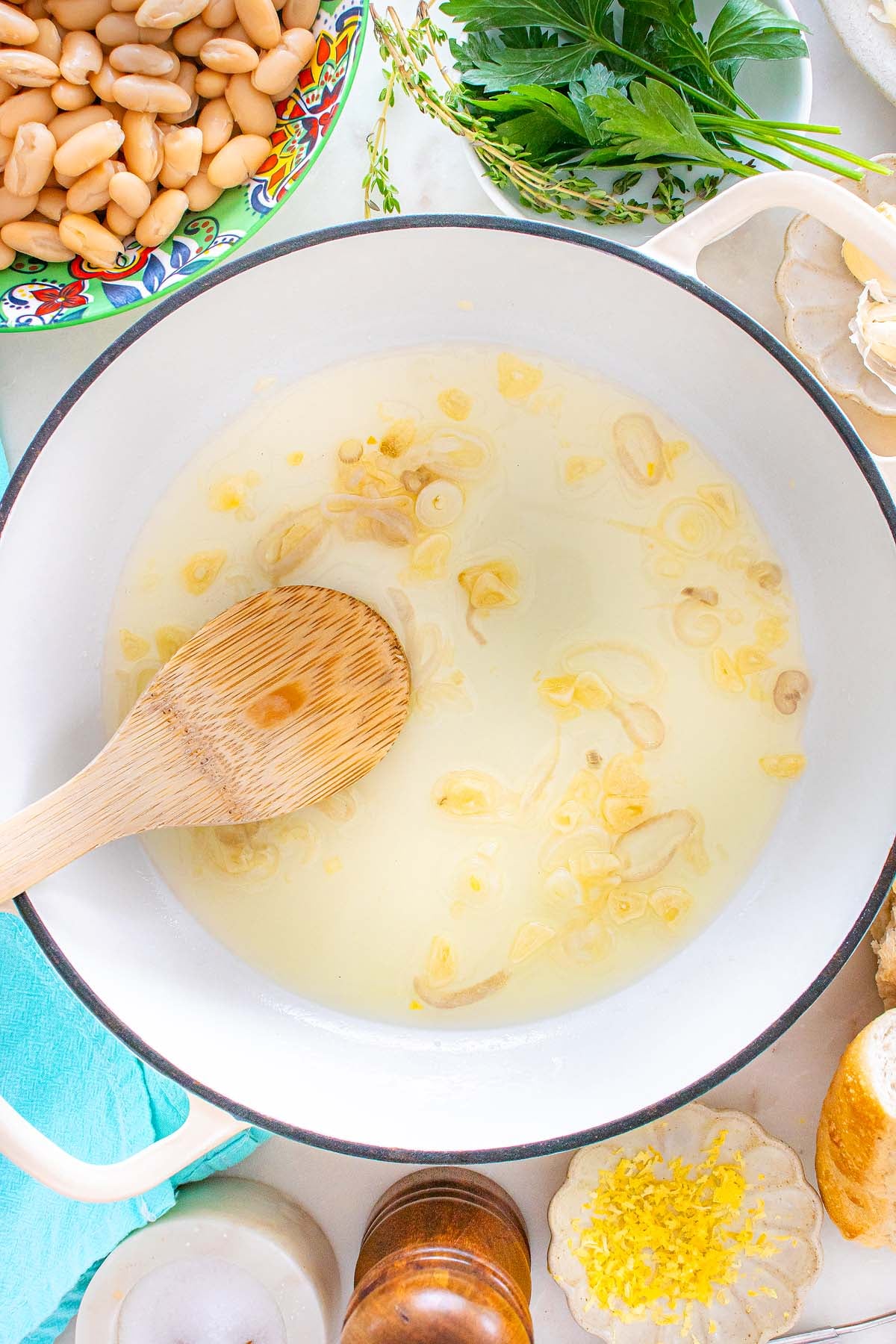 A wooden spoon stirs sliced garlic sautéing in oil in a white pot, surrounded by fresh herbs and Tuscan white beans.